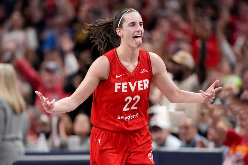 A close-up shot of Caitlin Clark wearing her red Indiana Fever jersey with the number 22, with her tongue sticking out in celebration and arms spread wide, on a basketball court with blurred crowd in the background.