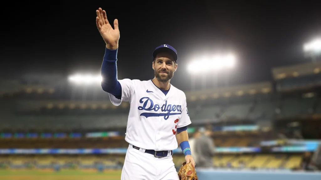Chris Taylor, a male baseball player in a white Los Angeles Dodgers uniform with a blue cap, waves to the crowd on a baseball field at night. He holds a brown baseball glove in his left hand and smiles, with stadium lights visible in the blurred background.
