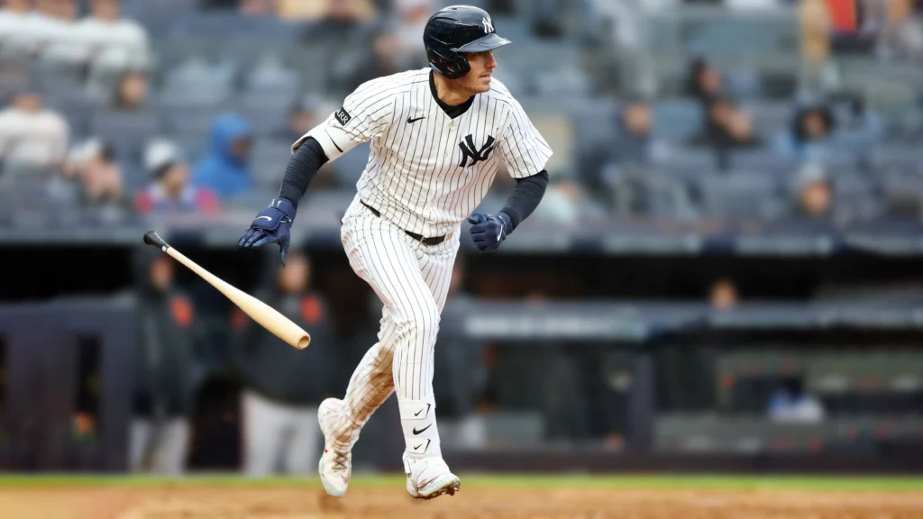 A baseball player wearing a white pinstriped New York Yankees uniform, a black helmet with the NY logo, and blue batting gloves is running towards first base on a baseball field. His bat is visible in the air behind him. The background shows blurred spectators in the stands.