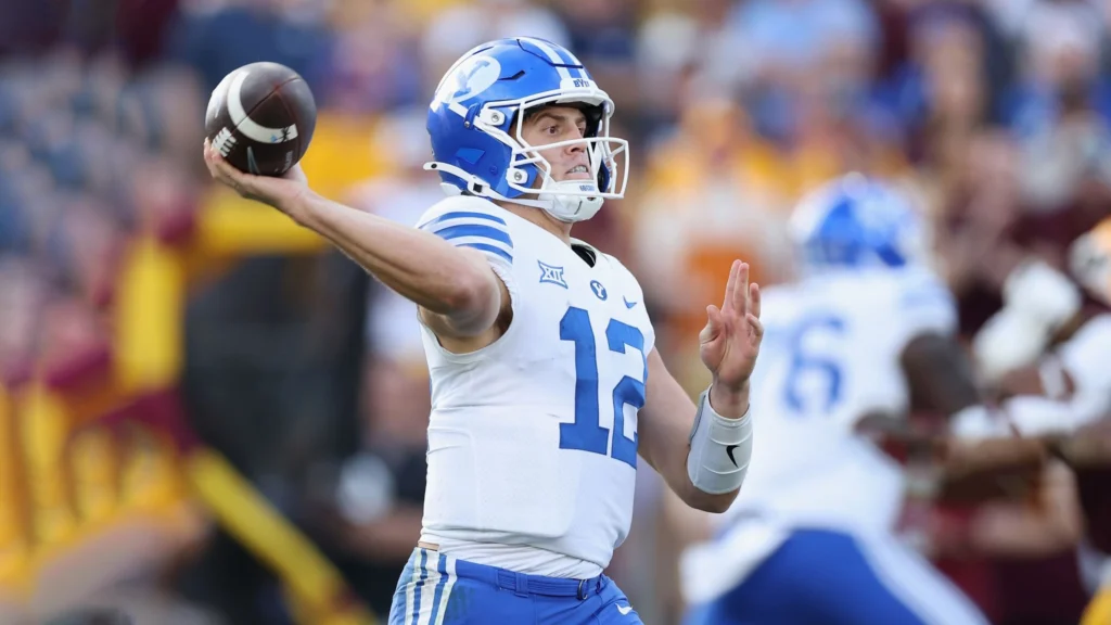 A football player, Jake Retzlaff, in a blue and white uniform with the number 16, is throwing a football. He is wearing a helmet and is on a football field.