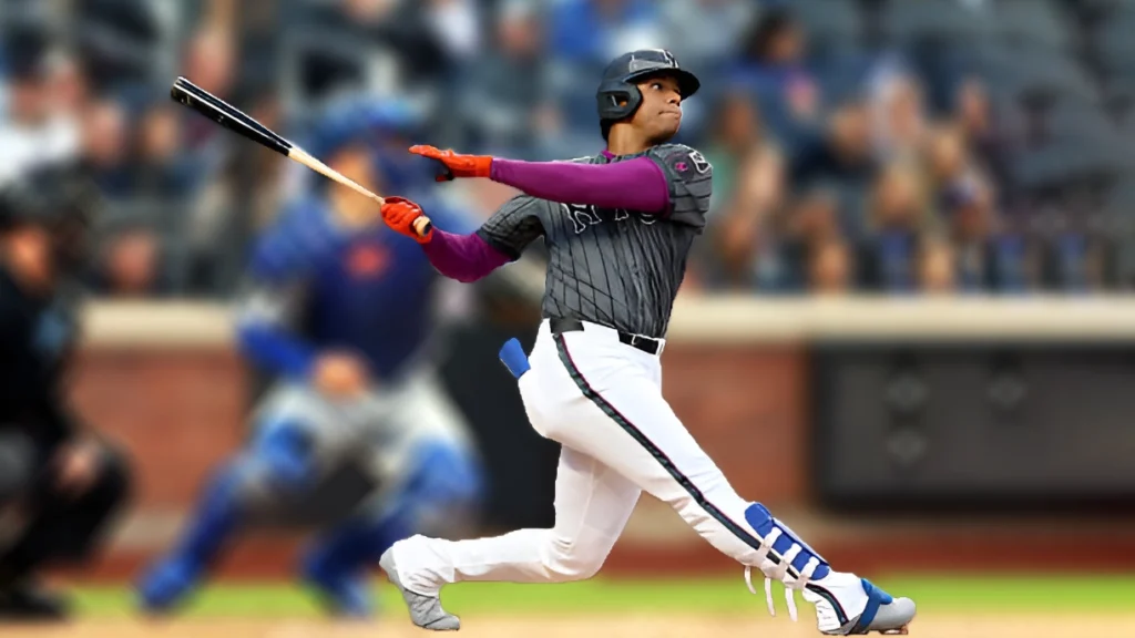 Baseball player Juan Soto, wearing a grey jersey with purple sleeves, white pants, and a helmet, swings a bat at a baseball. He is captured mid-swing with a focused expression, and the background is blurred with spectators.