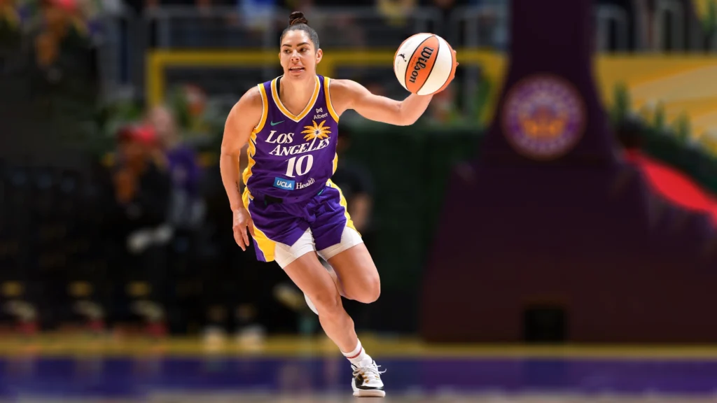 A female basketball player, Kelsey Plum, wearing a purple and gold Los Angeles Sparks jersey with the number 10, dribbles a Wilson basketball. She is looking forward with a determined expression as she moves across the court.