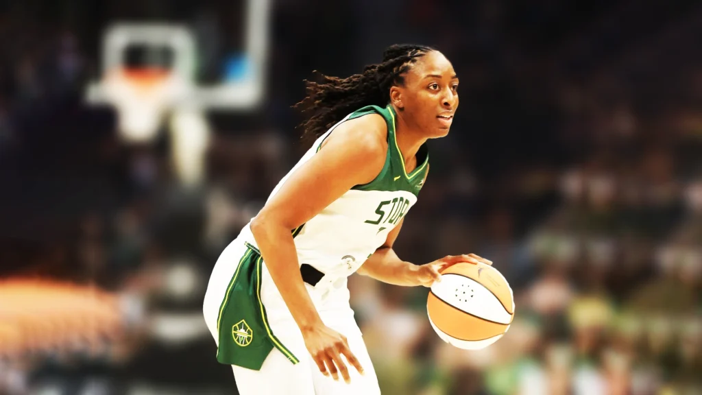 A full-body shot of Nneka Ogwumike, a Black woman with braided hair, on a basketball court. She is looking to her right while dribbling a basketball with her right hand. She is wearing a white and green Seattle Storm jersey and white shorts. The background is blurred, showing the arena and spectators.
