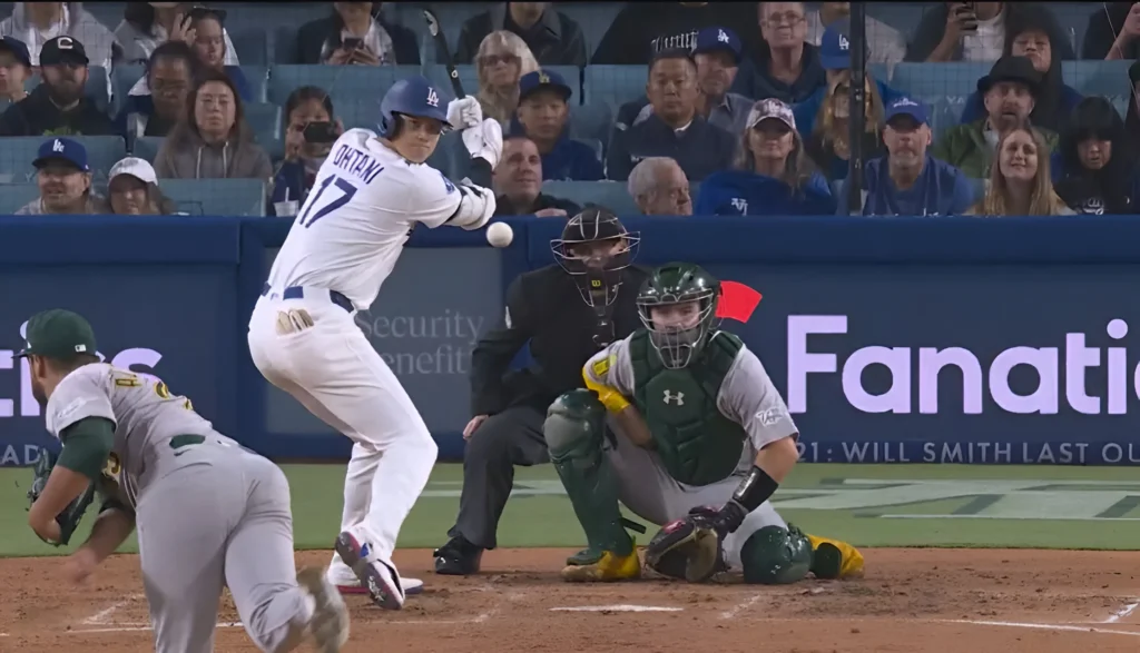 Baseball player Shohei Ohtani, wearing a white Dodgers uniform with the number 17, swings a bat at a baseball. A catcher in a green and grey uniform crouches behind home plate, and an umpire stands nearby. Spectators are visible in the background.