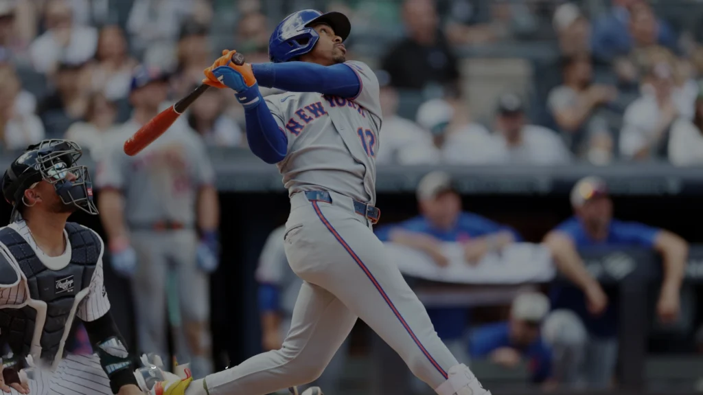 New York Mets shortstop Francisco Lindor wearing a blue helmet, gray jersey with "NEW YORK" across the chest, and gray pants with a blue stripe, swings a wooden baseball bat during a night game at Yankee Stadium. A New York Yankees catcher in a black helmet and pinstriped uniform crouches behind home plate. Spectators are visible in the blurred background.