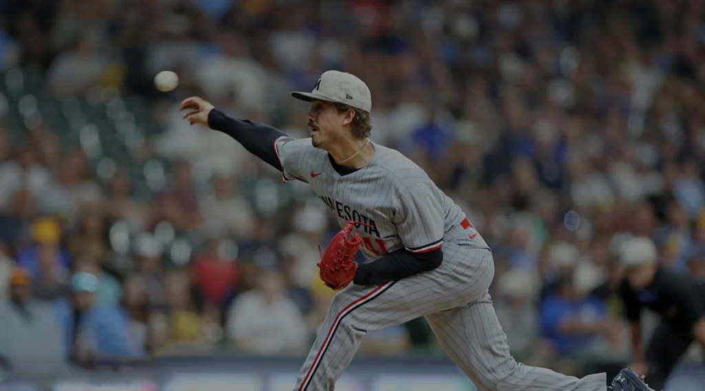 A baseball pitcher, Joe Ryan of the Minnesota Twins, in a gray uniform with pinstripes and a gray cap with a white "M" logo, is in the motion of throwing a baseball with his right hand. He has a red glove on his left hand and is positioned on a baseball field with a blurred background of spectators.