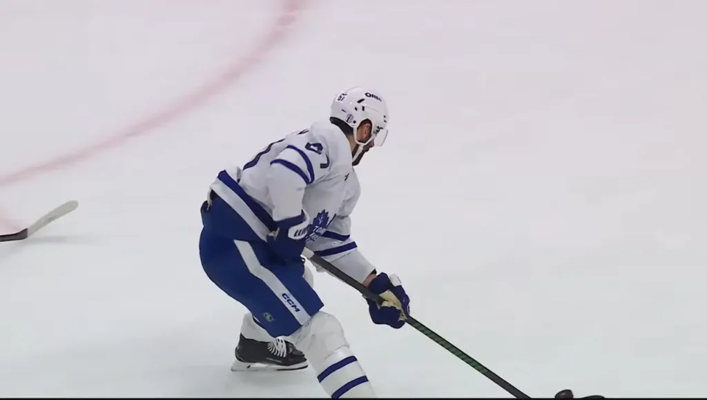 A hockey player, identified as Auston Matthews, wearing a white and blue Toronto Maple Leafs jersey, skates on the ice with his stick.