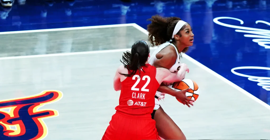 Caitlin Clark (jersey number 22) in a red Indiana Fever uniform pushing against Angel Reese who is holding a basketball. The court is blue and white, with team logos visible.