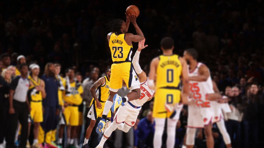 Aaron Nesmith, wearing a yellow Pacers jersey with the number 23, shoots a basketball while a Knicks player in a white jersey falls backward beneath him. Other basketball players and spectators are visible in the blurred background.