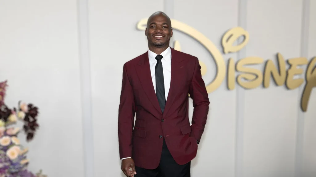 Adrian Peterson, a Black man in a burgundy suit and black tie, smiles widely at a Disney event. The gold "Disney+" logo is visible in the background.