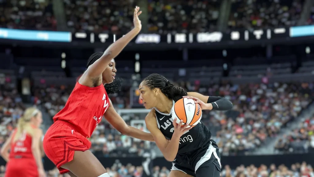 A'ja Wilson of the Las Vegas Aces in a black and white uniform dribbles the basketball while being defended by an opposing player in a red jersey of Fever, on a basketball court with a blurred crowd in the background.