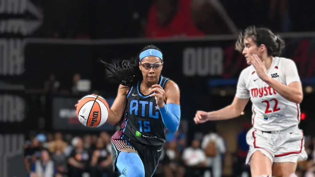 Allisha Gray, wearing an Atlanta Dream jersey with number 15, dribbles a basketball aggressively towards the basket. She is wearing a blue headband and arm sleeve. A Washington Mystics player in a white jersey with number 22 is guarding her, looking towards Gray. The game is taking place on a dark basketball court with blurred fans in the background.