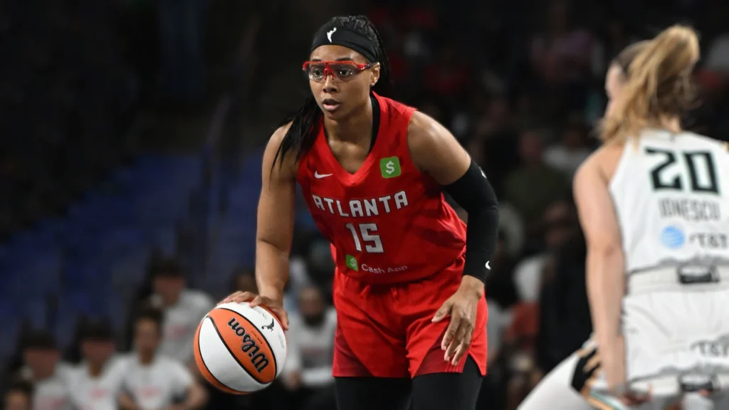 Allisha Gray, wearing a red Atlanta Dream jersey with the number 15 and protective goggles, dribbles a Wilson basketball on the court during a WNBA game. A New York Liberty player with the number 20 and "Ionescu" on her jersey is partially visible in the background.