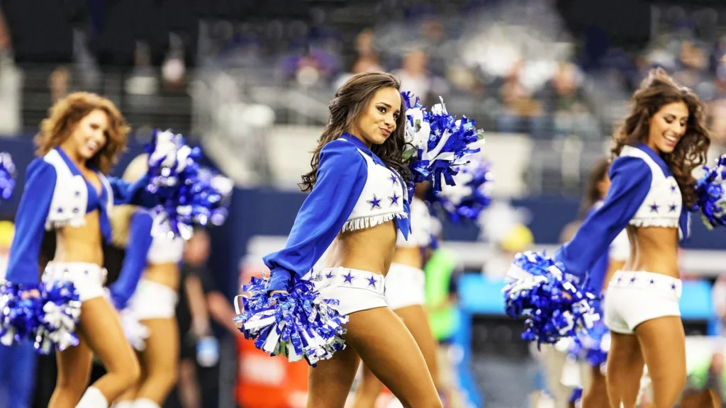 Three Dallas Cowboys Cheerleaders in their signature blue and white star-spangled uniforms perform on a football field, holding blue and white pom-poms. The cheerleader in the center smiles at the camera.