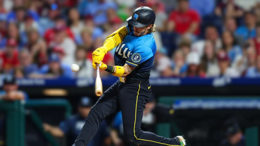 A male baseball player, identified as Bryson Stott by his uniform and previous context, in a blue and yellow jersey and black helmet, swings a wooden bat at a baseball in mid-air. He is in mid-swing with his body contorted in a powerful motion, wearing yellow sleeves and black pants. The baseball is visible near the end of his bat, slightly blurred, indicating motion. The background is a blurred stadium crowd, with hints of red and white, suggesting the team colors of the Philadelphia Phillies.