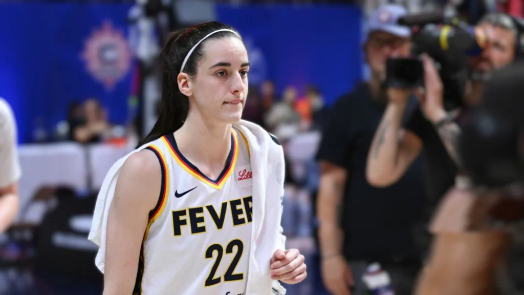 Caitlin Clark, an athlete, walks on a basketball court wearing a white Indiana Fever jersey with the number 22.
