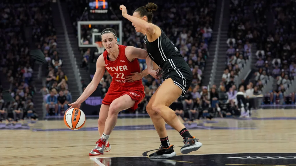 A basketball player in a red "Fever 22" jersey (Caitlin Clark) dribbles the ball with her left hand while being closely defended by an opponent in a black jersey. Both players are on a basketball court with blurred arena seating in the background during a game.