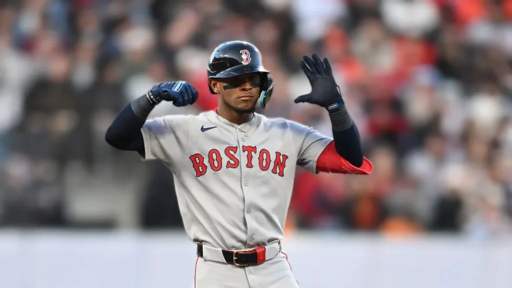 Ceddanne Rafaela, wearing a gray Boston Red Sox baseball uniform and helmet, raises his gloved hands in celebration on the field during a game. He is looking towards the left side of the frame with a focused expression. Spectators are visible in the blurred background.