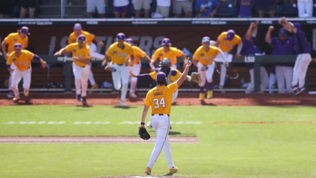 A baseball player, wearing an LSU uniform with "SHORES 34" on the back, stands on a pitcher's mound with one arm raised in celebration. Behind him, several teammates in yellow and purple uniforms run towards him from the dugout.