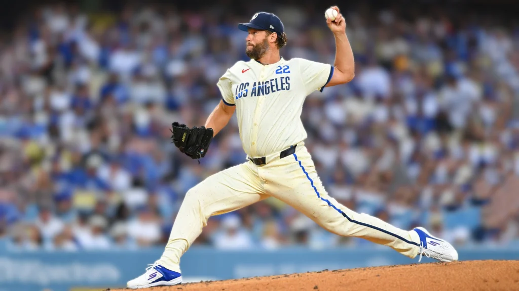 Clayton Kershaw, wearing a cream-colored Los Angeles Dodgers uniform with blue lettering and trim, is captured in mid-pitch motion on the mound at Dodger Stadium. He holds a baseball in his raised right hand, mid-release, with his left hand in a black baseball glove.