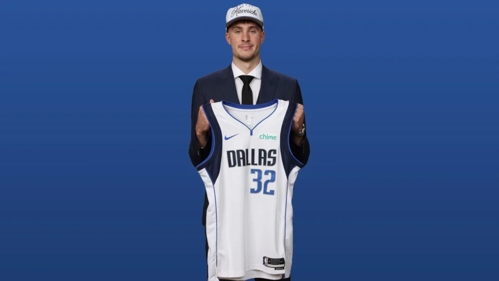 A young man, Cooper Flagg, standing against a blue background, wearing a dark suit, white shirt, and black tie. He is holding up a white Dallas Mavericks basketball jersey with "DALLAS" written across the chest in blue and the number "32" below it. He is also wearing a white baseball cap with "Mavericks" embroidered on it. He is looking directly at the camera with a slight smile.