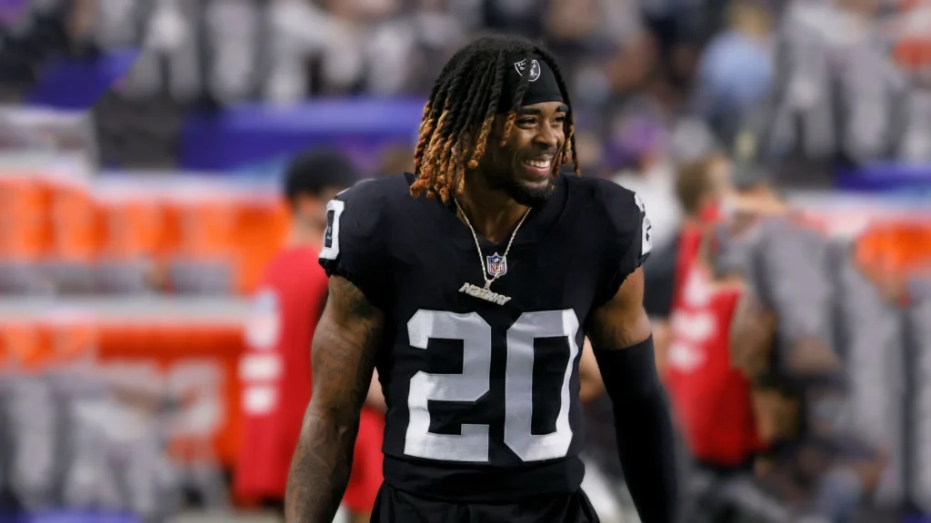 Close-up of Damon Arnette, a Black football player with dreadlocks, wearing a black Las Vegas Raiders jersey with the number 20, smiling on a football field.