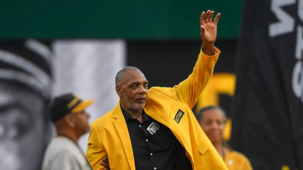 A middle-aged Black man with a graying beard and short dark hair, wearing a bright yellow jacket over a black shirt, waves with his right hand raised. He is looking slightly to his left. In the background, out of focus, are parts of other people and a green and black banner.