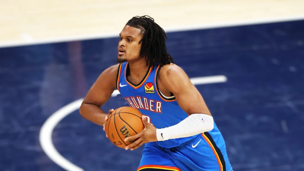 Dillon Jones, a male basketball player with dreadlocks, in an Oklahoma City Thunder blue and orange jersey, dribbling a basketball on a court. He is looking to his left, appearing focused during a game.