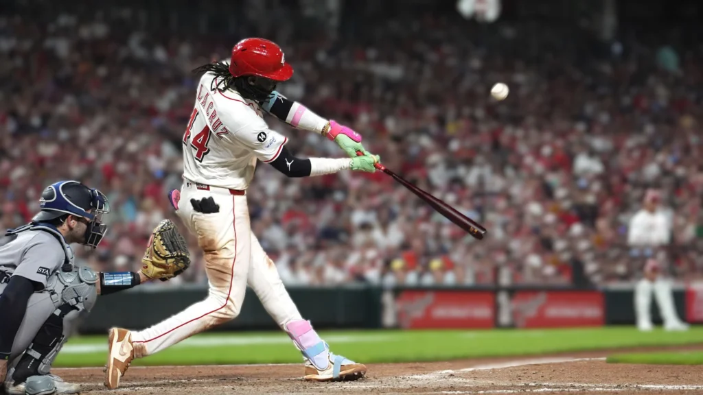 Elly De La Cruz, a baseball player in a red and white uniform, swings a bat at a baseball during a game.