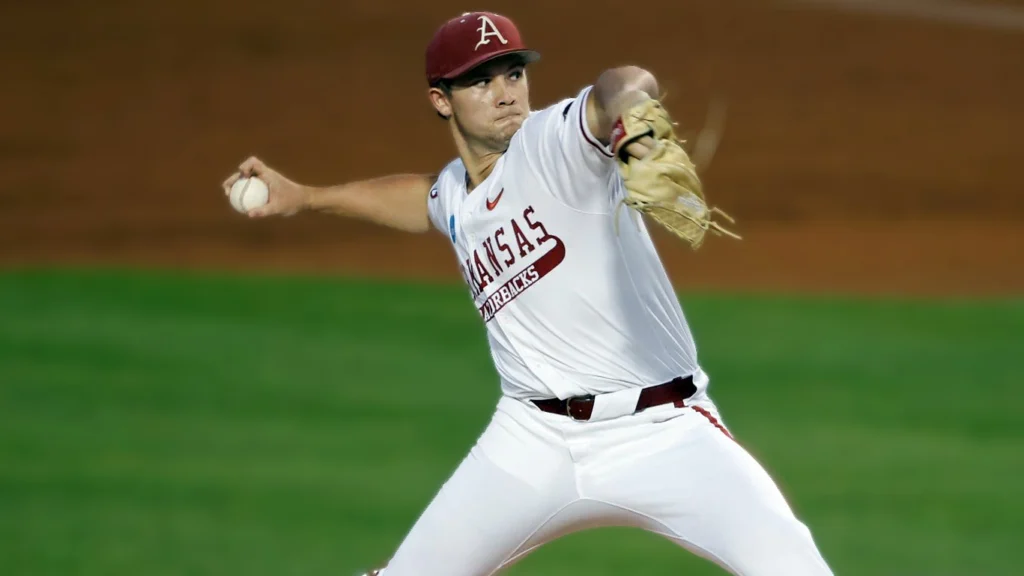 A male baseball pitcher in a white Arkansas Razorbacks uniform with a red cap and glove, mid-throw on a baseball mound with green grass in the background.