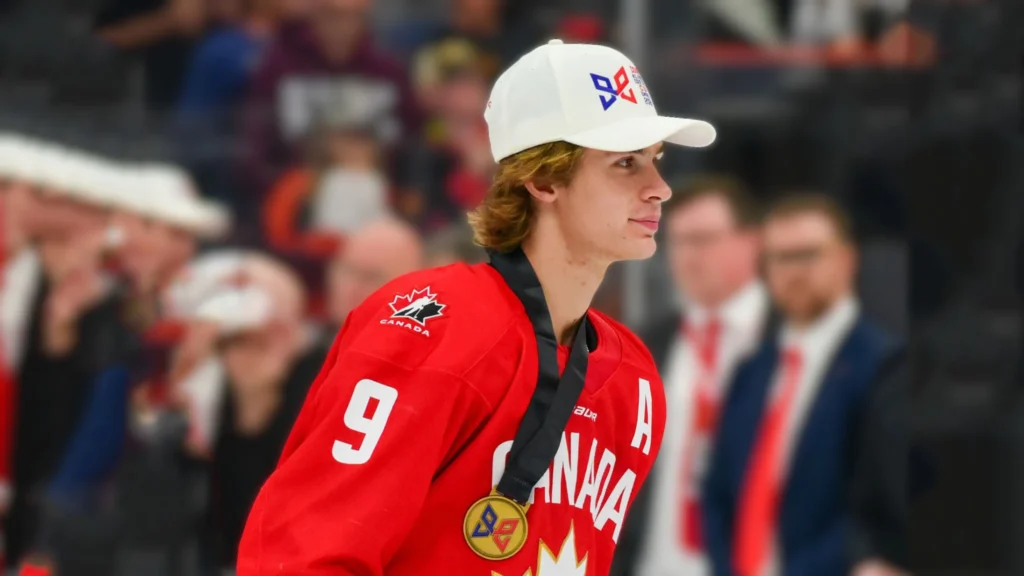 A young male hockey player, Gavin McKenna, with wavy blond hair, wearing a red Team Canada jersey with the number 9, a gold medal around his neck, and a white baseball cap. He is looking to his right with a slight smile. The background is blurred, showing spectators.