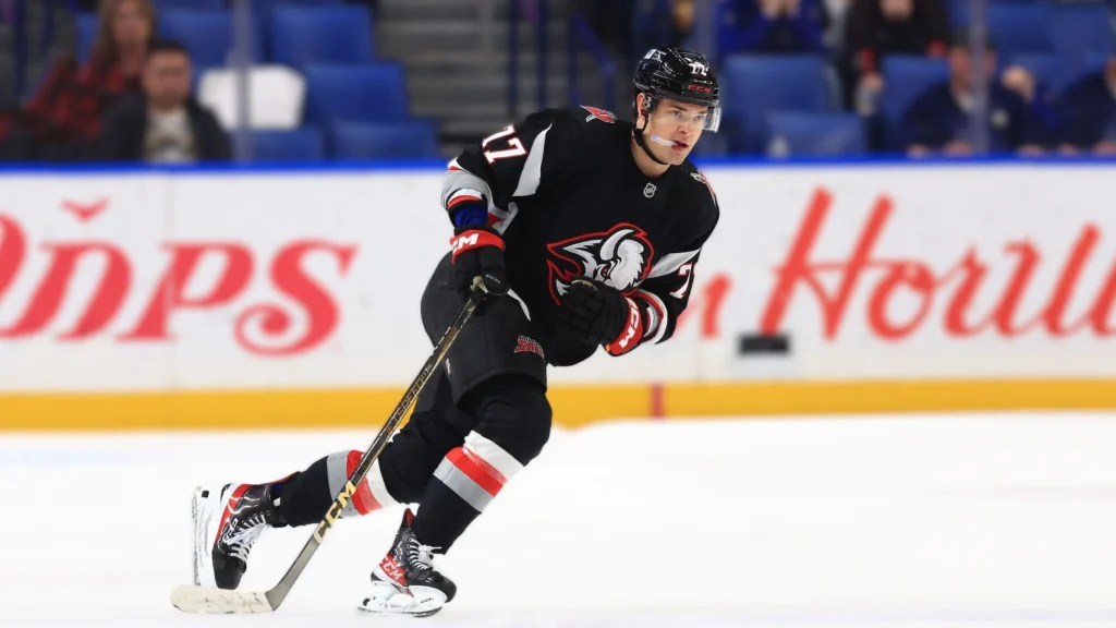 A full-length shot of NHL player JJ Peterka, wearing a black Buffalo Sabres jersey with the number 77, skating on the ice with a puck on his stick during a game. He is looking forward, and his posture shows speed and focus.