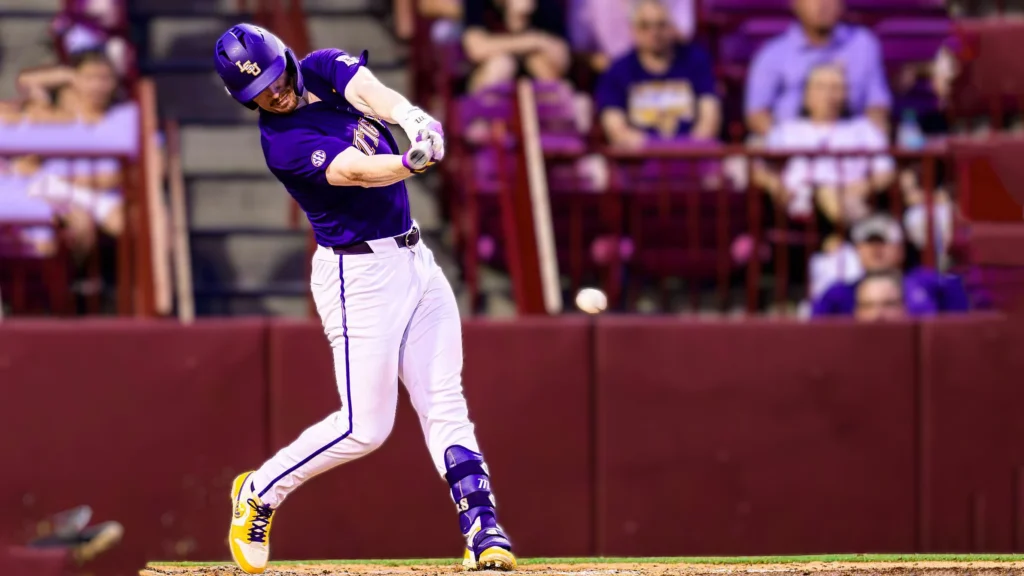 LSU baseball player Jared Jones in a purple and white uniform mid-swing, hitting a baseball. The ball is visible in the air near the bat.