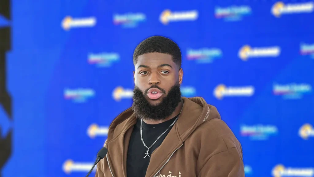 Portrait of NFL player Jared Verse wearing a brown hoodie and chain, speaking at a microphone with a blue background, in the context of his recent comments about the Philadelphia Eagles fan base.