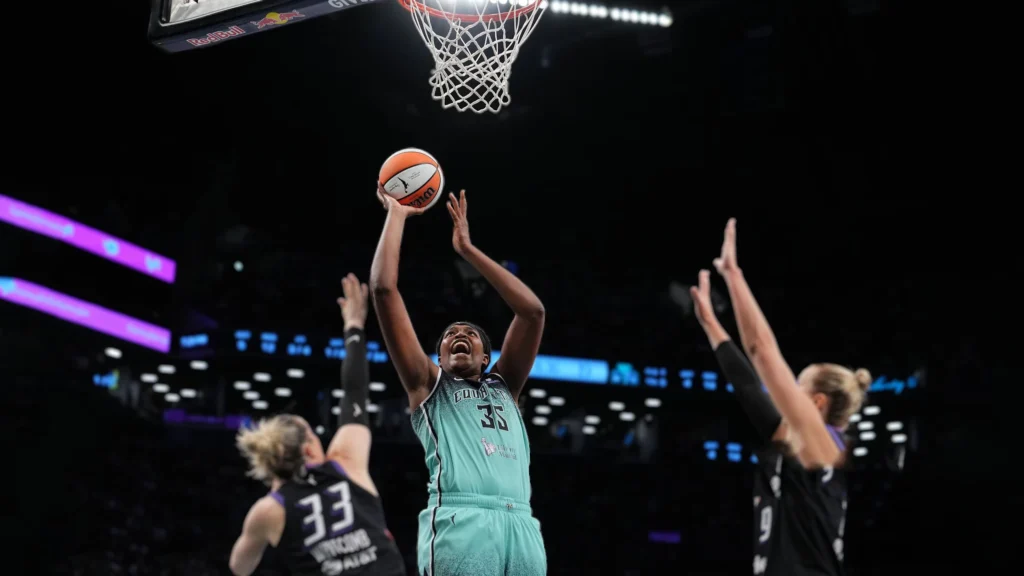 A basketball player in a teal jersey with "35" on it, identified as Jonquel Jones, extends her arms towards a basketball hoop, about to shoot. Two opposing players in dark jerseys are attempting to defend her. The background shows a dimly lit arena crowd.Jones went down in the second quarter after an awkward landing on a drive to the basket, colliding with Mercury defender Sami Whitcomb.
