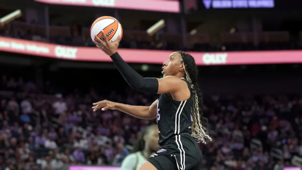 Golden State Valkyries player Kayla Thornton in a black jersey with white stripes and braided hair, driving towards the basket with a basketball in her right hand during a WNBA game. A blurry crowd is in the background.