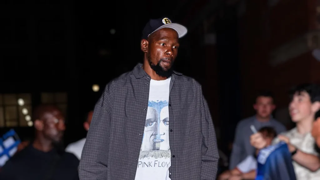 Kevin Durant, a tall male athlete with a beard, wearing a baseball cap, a grey plaid jacket over a white t-shirt with a Pink Floyd graphic, standing in a dimly lit setting with people in the background.