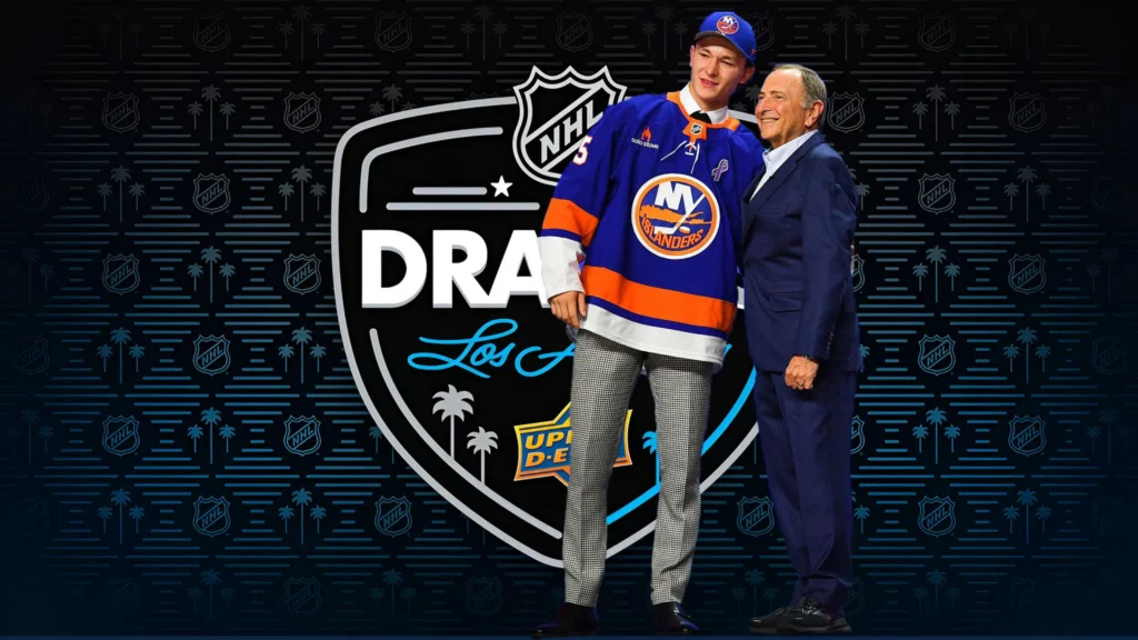 A tall, smiling young man Matthew Schaefer, in a New York Islanders jersey and a baseball cap stands next to a smiling man in a suit, NHL Commissioner Gary Bettman, on the draft stage. The backdrop is dark with a logo for the NHL Draft in Los Angeles.
