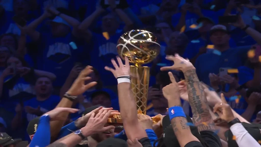 A close-up shot of multiple hands, some tattooed, reaching up to touch and hold the gold Larry O'Brien NBA Championship Trophy. The trophy is gleaming in the center, surrounded by a celebratory crowd in blue jerseys, indicating the Oklahoma City Thunder team and their fans.