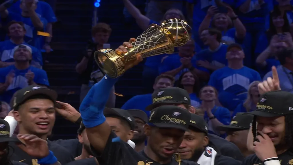 Shai Gilgeous-Alexander, wearing an Oklahoma City Thunder jersey and a "Champions" hat, holds the golden NBA Finals MVP trophy above his head, celebrating with his teammates on the court as cheering fans are visible in the background.