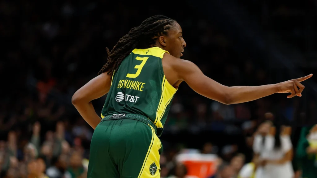 Nneka Ogwumike, wearing a green Seattle Storm jersey with the number 3 and her name on the back, stands on a basketball court, looking over her right shoulder and pointing forward with her left hand, appearing to direct a play.