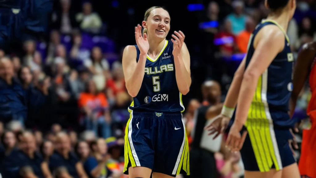 Paige Bueckers, wearing a dark blue Dallas Wings jersey with the number 5, smiles and claps her hands together on a basketball court. Her blonde hair is pulled back, and she is looking towards her right. Other players and blurred spectators are visible in the background.
