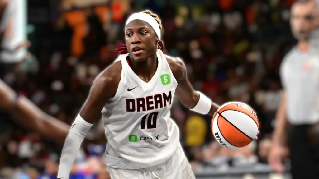A close-up action shot of WNBA player Rhyne Howard of the Atlanta Dream, wearing a white jersey with "DREAM" and number 10, dribbling a basketball with intense focus during a game. She has a white headband and braided hair.