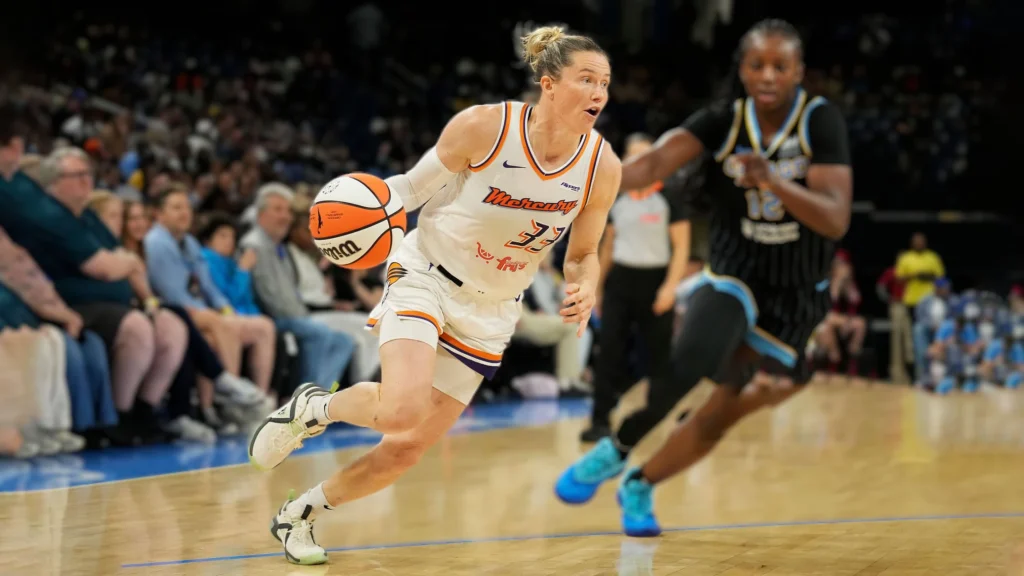 Phoenix Mercury guard Sami Whitcomb dribbles the basketball downcourt during a WNBA game against the Chicago Sky. She is wearing a white and orange Mercury uniform with number 33. A Chicago Sky player in a black and blue uniform defends closely behind her.