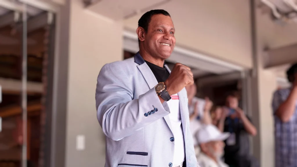 A smiling Sammy Sosa, wearing a light blue patterned suit jacket and a dark shirt, stands in a stadium box with his right arm raised in a triumphant or greeting gesture. He is looking off to his right with a happy expression, and a crowd can be faintly seen in the background.