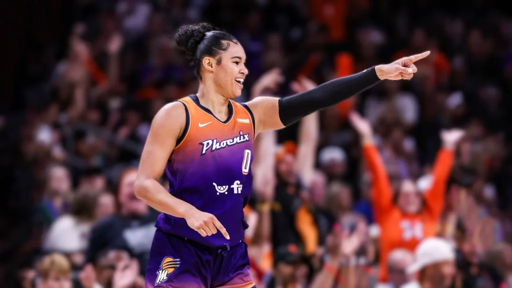 Satou Sabally, a Phoenix Mercury basketball player, in her purple and orange jersey, smiling and pointing while on the court during a game with fans visible in the background.