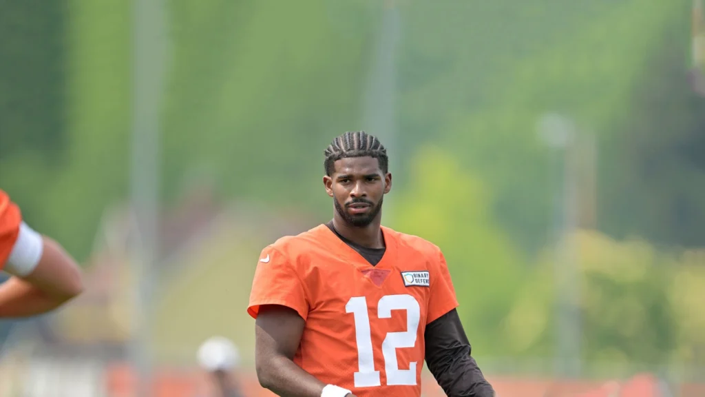 Shedeur Sanders, wearing an orange Cleveland Browns practice jersey with the number 12, looks directly at the camera on a sunny day with green trees in the blurry background.