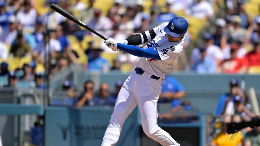 Shohei Ohtani, wearing a white Dodgers uniform and blue helmet, in mid-swing during a baseball game with fans visible in the stands behind him.