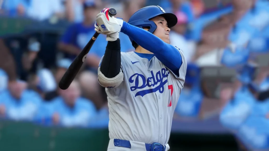 Shohei Ohtani in a Los Angeles Dodgers uniform, mid-follow-through after hitting the ball, with a blurred baseball stadium crowd in the background during a game.
