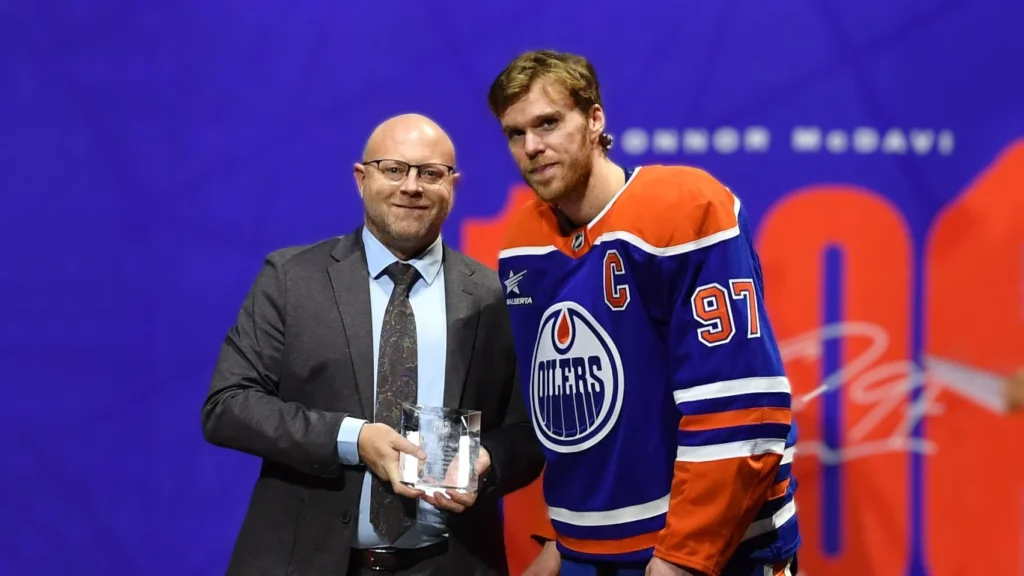 Connor McDavid in an Edmonton Oilers jersey with the captain's "C" and number 97, standing next to a bald man in a suit, who is holding a clear award. They are on a stage with a blue background and large orange graphics that partially show "CONNOR MCDAVID" and "97"